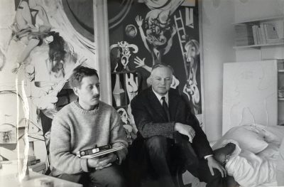 Zwei Männer sitzen nebeneinander. Ihr Blick ist nach unten gerichtet. Im Hintergrund hängt ein Gemälde an der Wand.
