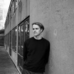 Schwarz-weiß Fotografie. Jan Sagasser lehnt an einer Außenwand der HfG Ulm und blickt zur Seite.