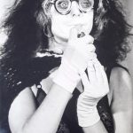 Schwarz-weiß -Fotografie: Porträt einer langhaarigen Frau. Sie trägt eine Brille und weiße Handschuhe.