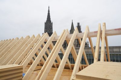 Farbfoto: Blick auf Holzlatten in einem Dachstuhl, ein Mann geht hindurch. Im Hintergrund vor grauem Himmel das Ulmer Münster