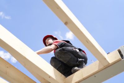 Detailaufnahme eines Bauarbeiters mit Helm zwischen zwei hellen Holzlatten des Dachstuhls vor blauem Himmel