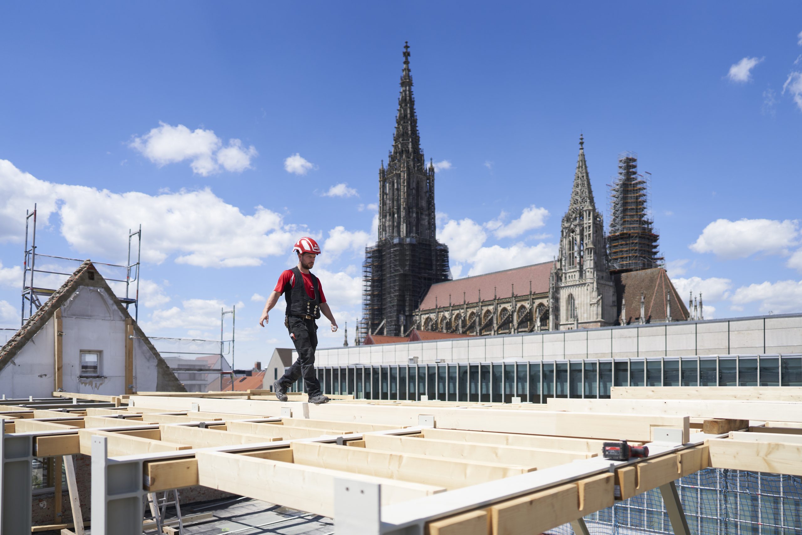 Farbfoto: Blick auf Holzlatten in einem Dachstuhl, ein Mann geht hindurch. Im Hintergrund vor blauem Himmel das Ulmer Münster