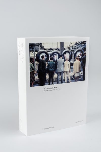 Farbfoto: Ein weißes Buch steht aufgestellt auf einem hellgrauen Tisch und Hintergrund