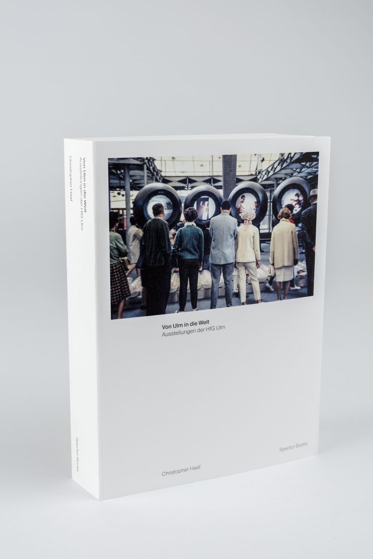 Farbfoto: Ein weißes Buch steht aufgestellt auf einem hellgrauen Tisch und Hintergrund