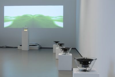 Farbfoto: Ansicht eines Ausstellungsraums mit weißen Wänden. An der hinteren Wand eine Videoprojektion, darunter auf einem weißen Sockel eine Glasschüssel und an den Seitenwänden rechts und links jeweils 4 Glasschüsseln mit Lautsprechern auf weißen Sockeln.