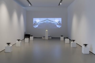 Farbfoto: Ansicht eines Ausstellungsraums mit weißen Wänden. An der hinteren Wand eine Videoprojektion, darunter auf einem weißen Sockel eine Glassschüssel und an den Seitenwänden rechts und links jeweils 4 Glasschüsseln mit Lautsprechern auf weißen Sockeln.