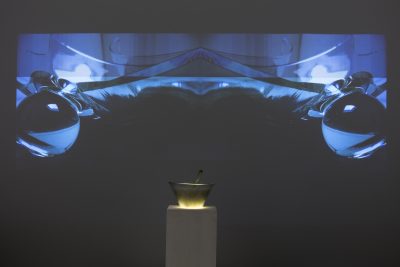 Farbfoto: Ansicht eines Ausstellungsraums mit weißen Wänden. An der hinteren Wand eine Videoprojektion, darunter auf einem weißen Sockel eine Glasschüssel und an den Seitenwänden rechts und links jeweils 4 Glasschüsseln mit Lautsprechern auf weißen Sockeln.