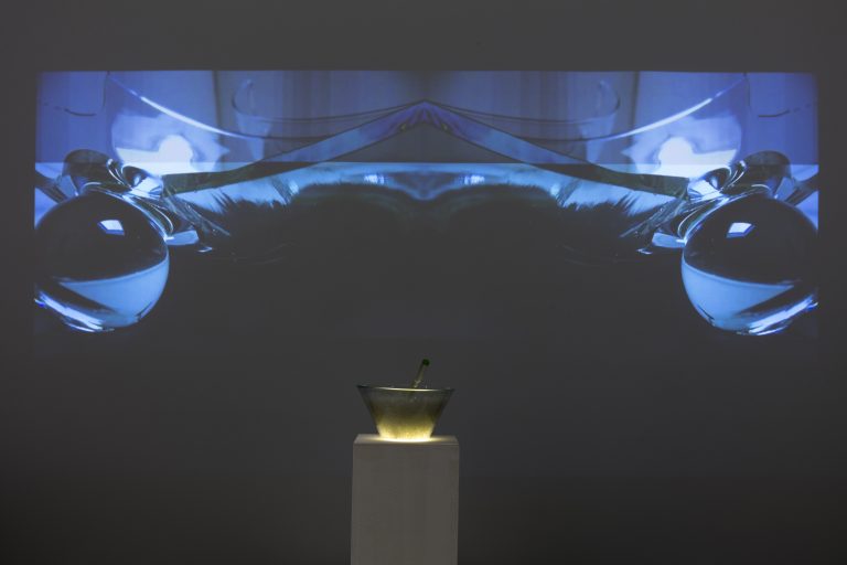 Farbfoto: Ansicht eines Ausstellungsraums mit weißen Wänden. An der hinteren Wand eine Videoprojektion, darunter auf einem weißen Sockel eine Glassschüssel