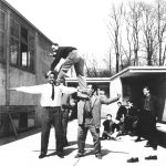Schwarz-Weiß-Fotografie: Vor einem flachen gebäude aus Beton stehen mehrere junge Männer in Anzügen. Einer ist auf die Schultern von beiden anderen geklettert. Sie schauen lachend in die Kamera.